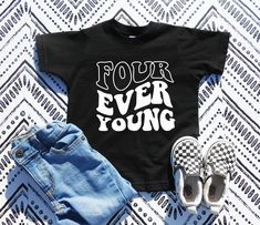 the young wild and three shirt is laying on top of some denim shorts, with shoes next to it