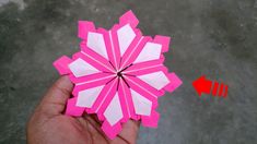 a hand holding a pink and white origami snowflake with an arrow pointing to it