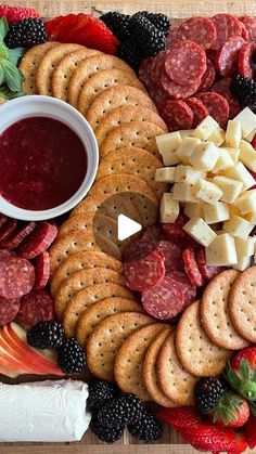 a platter filled with cheese, crackers, fruit and sausage next to a bowl of jam