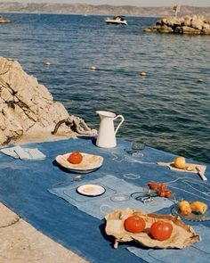 a picnic table with plates and fruit on it near the water's edge,