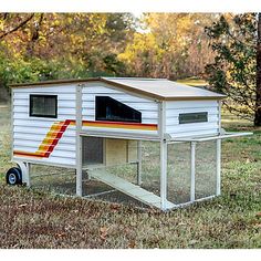 a white chicken coop with a red and yellow stripe on the side, sitting in grass next to trees