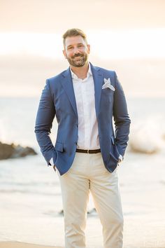 a man in a blue jacket and white shirt standing on the beach with his hands in his pockets