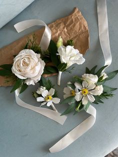 three white flowers with green leaves and ribbons on a blue table cloth next to a piece of brown paper