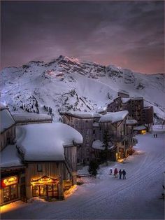 people are walking in the snow near buildings and mountains at night with lights shining on them