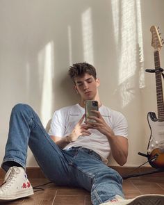 a young man sitting on the floor looking at his cell phone while playing guitar in front of him