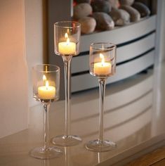 three wine glasses with lit candles in them on a glass table next to a fireplace