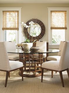 a dining room table with chairs and a round mirror on the wall above it in front of two windows