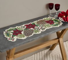 a decorative table runner with poinsettis and pine cones on it next to two wine glasses