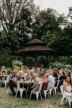 a group of people sitting around a wooden table in the middle of a field with white chairs