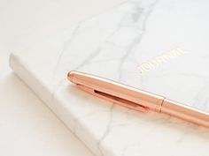 a rose gold pen sitting on top of a white marble book with the word journal written in gold