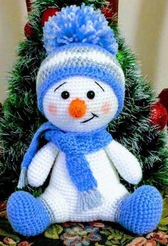 a crocheted snowman sitting in front of a christmas tree with a blue hat and scarf