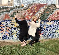 two women standing in front of a wall with mosaic tiles on it and one holding up her hand