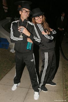 two people dressed in black standing next to each other on a sidewalk at night time