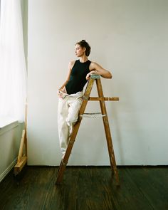 a woman sitting on top of a wooden ladder next to a white wall and window