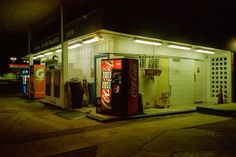 an old gas station at night with the lights on and soda machines in front of it