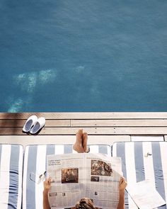 a person sitting on top of a deck reading a newspaper next to the ocean with their feet up