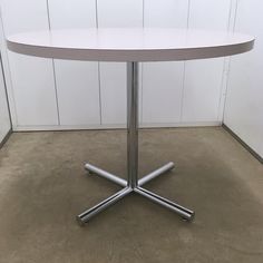 a white table with four silver legs on concrete flooring in front of a white wall