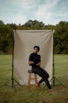 a woman sitting on top of a stool in front of a white backdrop with trees