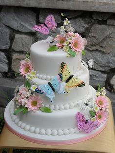 a three tiered cake with butterflies and flowers on the top, sitting on a table in front of a stone wall