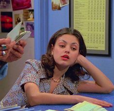 a woman sitting at a table with her hand on her head and another person holding a cell phone to her ear