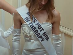 a woman in a white dress and gloves is wearing a sash with words on it