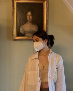 a woman wearing a face mask standing in front of a portrait on the wall with a painting behind her