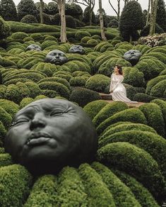 a woman sitting on top of a lush green field next to trees and bushes in the shape of heads