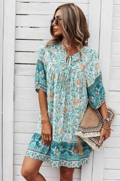 A woman in sunglasses stands against a white wooden wall, wearing the trendy Ruston Floral Boho Dress from Western Edge Boutique in floral turquoise and beige. She holds a woven clutch with tassels, her wavy hair flowing as she looks off to the side on this perfect summer day. Chic V-neck Maxi Dress For Summer Outings, Casual Knee-length Boho Print Dress, Bohemian Floral Print Knee-length Sundress, Casual Boho Dress With Knee-length Boho Print, Bohemian Knee-length Floral Sundress, Casual Boho Print Sundress For Beach Season, Knee-length Boho Print Summer Dress, Casual Knee-length Boho Dress With Boho Print, Knee-length Boho Print Dress For Summer