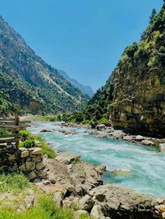 a river flowing through a lush green valley next to a rocky mountain side under a blue sky