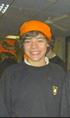 a young man wearing an orange hat and black shirt smiling at the camera with other people in the background