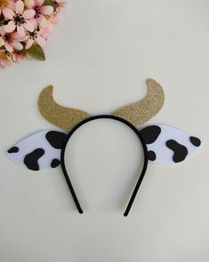 a cow ears headband with gold horns and black spots on it, sitting next to pink flowers