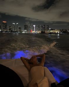 a man laying on the back of a boat at night with city lights in the background