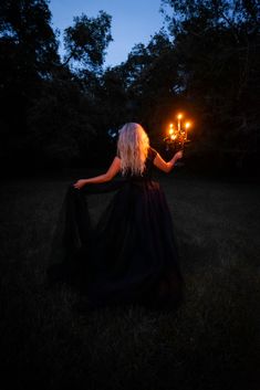 a woman in a long black dress holding two candles up to her face at night