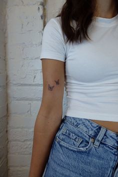 a woman with a small butterfly tattoo on her left arm, standing in front of a white brick wall