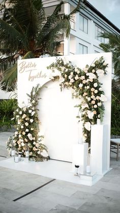 an outdoor ceremony setup with white flowers and greenery on the wall, surrounded by palm trees