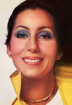 a woman with blue eyes and yellow jacket smiling at the camera while wearing gold jewelry