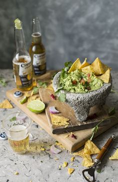 a wooden cutting board topped with guacamole and tortilla chips next to two bottles of beer