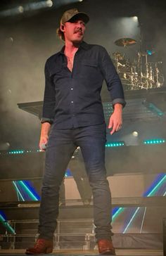 a man standing on top of a stage with his hands in his pockets and wearing a hat