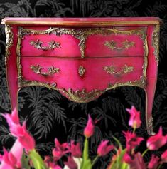 a pink and gold painted chest of drawers with flowers on the bottom, in front of a black wallpapered background