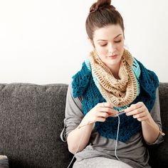 a woman sitting on a couch holding a crocheted scarf and knitting it with her hands