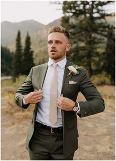 a man wearing a suit and tie standing in front of some trees with his hands on his hips