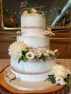 a three tiered wedding cake with flowers and greenery on the top is displayed in front of a mirror