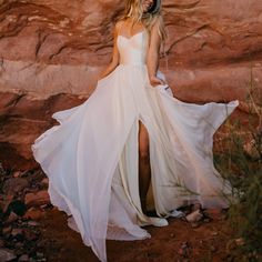 a woman wearing a white dress standing in front of a rock formation with her legs crossed