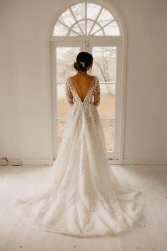 a woman standing in front of a window wearing a wedding dress with long sleeves and an open back