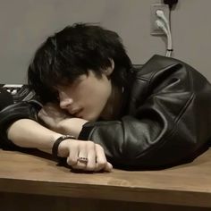 a young man wearing a black leather jacket leaning on a wooden table with his head resting on his hand