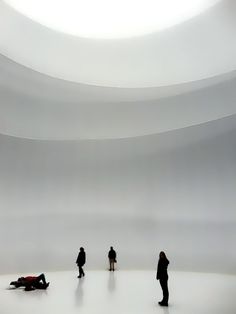 three people standing in front of a white wall with a circular light above them and one person laying on the floor