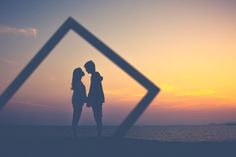 two people standing next to each other near the ocean at sunset with their reflection in the frame