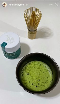 a green drink in a black bowl next to a whisk on a white table