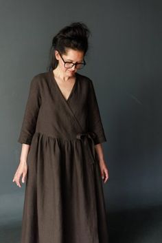 a woman standing in front of a gray wall wearing glasses and a dark brown dress