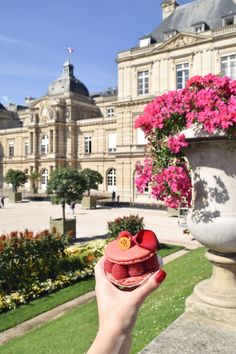 a person holding up a small red cupcake in front of a building with pink flowers